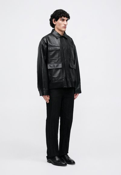 Uomo in piedi che indossa una giacca di pelle nera con diverse tasche, pantaloni neri testurizzati e scarpe di pelle nere su uno sfondo bianco semplice.