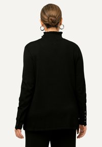 Pull noir avec un col haut à volants. Les manches longues présentent des détails de boutons sur les poignets. Tissu doux avec une texture lisse.