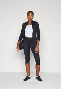 Melnas sporta jaka ar rāvējslēdzēju, balta topiņa un melnas kapri legingi. Valkāts kopā ar melnām sporta kurpēm un tur melnu somu.