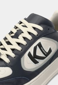 Αθλητικό παπούτσι σε μπλε ναυτικό και γκρι χρώμα με κρεμ κορδόνια, κατασκευασμένο από δέρμα και σουέτ, με ανυψωμένο μαύρο λογότυπο και διάτρητο σχέδιο στο πλάι.