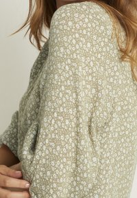 Bluse mit Blumenmuster aus olivgrünem Stoff, verziert mit zarten weißen Blumen, kurzen Ärmeln und weicher Textur. Nahaufnahme des Ärmeldetails.