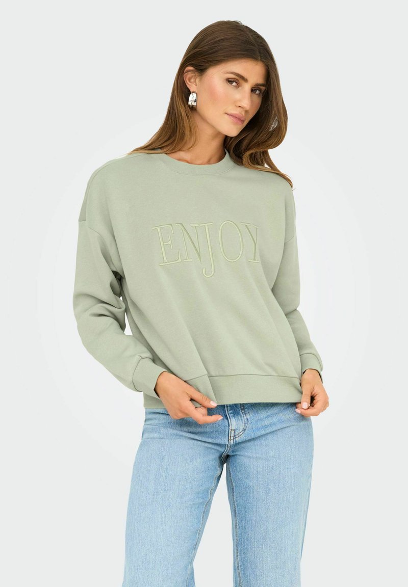Vrouw in een saliegroen sweatshirt met de tekst "ENJOY", lichtblauwe jeans en zilveren oorbellen, staand tegen een effen achtergrond.