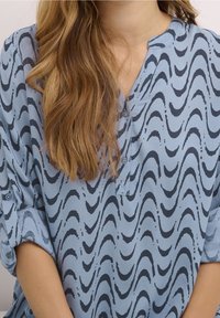 Niebieska bluzka z falistym czarnym wzorem, lekkiego materiału, z krótkimi rękawami i dekoltem w szpic z guzikami.