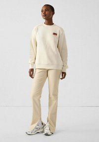 Sudadera crema con un logotipo rojo y negro, combinada con pantalones divididos beige. El modelo lleva zapatillas deportivas grises. Textura suave, ajuste relajado.