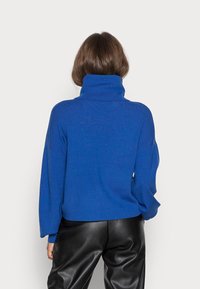 Pull à col roulé bleu avec une texture côtelée, des manches larges et une coupe décontractée, associé à un pantalon en cuir noir.
