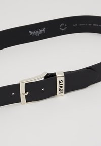 Ceinture en cuir noir avec une boucle en métal argenté, présentant une brandissage en relief. Texture lisse et forme rectangulaire classique.