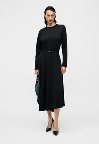 Μακρύ μαύρο φόρεμα με στρογγυλή λαιμόκοψη, μακριά μανίκια και ζώνη στη μέση. Φορεμένο με μαύρες ψηλοτάκουνες γόβες και πράσινη τσάντα. Λεία υφή υφάσματος.