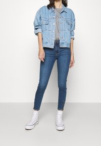 Ljusblå denimjacka över en grå t-shirt, kombinerad med mörkblå skinny jeans och vita höga sneakers. Släta texturer och avslappnad passform.