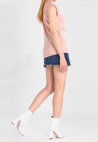 Světle růžový top s krátkým rukávem a krajkovými detaily, doplněný o džínové šortky. Modelka má na sobě bílé holínky na podpatku se zipem a béžovou podrážkou.