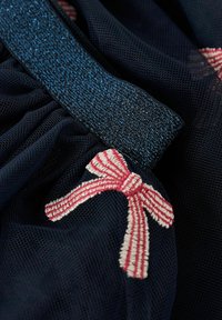Marinblå nättextil med rynkad struktur och ett glittrande blått elastiskt band. Har en röd och vit randig broderad båge som accent.