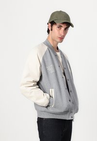 Mladý muž v šedé varsity bundě s krémovými rukávy, olivově zelené čepici a tmavých džínech, stojící proti jednoduchému bílému pozadí.