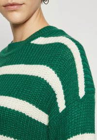Maglione a righe verdi e crema, che mostra un motivo testurizzato. Presenta un collo rotondo e una vestibilità morbida, con dettagli di cucitura visibili.