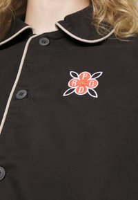 Fekete gombos ing, fehér díszítéssel, amelyen egy piros és fehér hímzett virág található a mellrészen, P, R, D és G betűkkel.