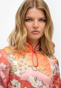 Blusa de seda con cuello mandarín. Estampado floral en tonos rosa y rojo con acentos naranjas, con detalle de ojo de cerradura en el escote.