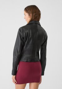 Giubbotto in pelle nera con un design aderente, colletto a scannellature e dettagli di cucitura sottili. Indossato con una mini gonna bordeaux.