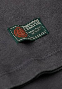 Tela de camiseta de algodón gris oscuro con una etiqueta cosida. La etiqueta presenta texto verde y naranja: "Superdry", "Vintage Athletic Outfitters."