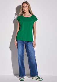 Vihreä lyhythihainen paita pyöreällä kauluksella ja olkapäillä laskokset, yhdistettynä sinisiin leveälahkeisiin farkkuihin ja vihreillä yksityiskohdilla varustettuihin lenkkareihin.