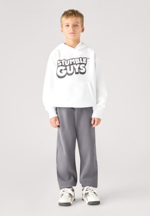Ragazzo con felpa bianca con cappuccio con il testo "Stumble Guys", pantaloni grigi e scarpe da ginnastica bianche, in piedi davanti a uno sfondo semplice.