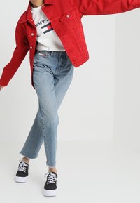 Veste en denim rouge sur une chemise blanche à rayures bleu et rouge, associée à un jean bleu clair et des baskets noires avec des semelles blanches.