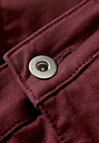 Close-up van bordeauxkleurige stof met een zilverkleurige metalen knoop met ingegraveerde merknaam, vergezeld van een zichtbare stikselstructuur.