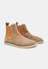 Bottes Chelsea en suède beige avec élastiques latéraux orange, tirant brun et semelle en caoutchouc beige. Texture lisse, forme de bout arrondi et détails cousus.