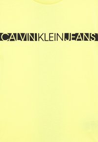 Žluté bavlněné tričko s černým grafickým potiskem "CALVIN KLEIN JEANS" přes hruď v tučném, moderním písmu.