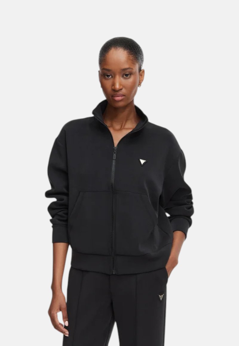 Jeune femme aux cheveux courts portant une veste zippée noire et un pantalon noir assorti avec un petit logo triangulaire blanc.