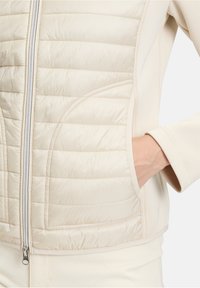 Beżowa puchowa kurtka o pikowanej fakturze, z zamkiem z przodu i bocznymi kieszeniami. Gładkie akcenty materiałowe na rękawach.