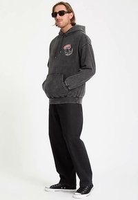 Grijze hoodie met voorzak, grafisch ontwerp op de borst, gecombineerd met zwarte losse broek en zwarte sneakers. Model draagt een zonnebril.