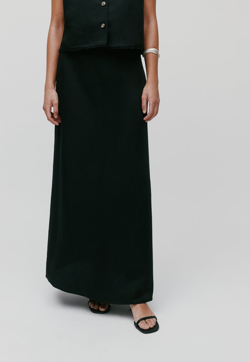Jupe maxi noire en tissu doux, offrant une texture lisse et une silhouette droite, associée à des sandales noires simples.