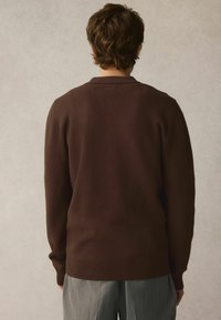 Brauner Strickpullover mit lockerer Passform und gerippten Bündchen, getragen über hellgrauen gestreiften Hosen. Der Rücken hat ein einfaches Kragen Design.