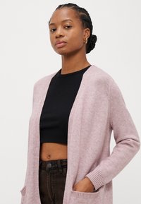 Világos rózsaszín kardigán nyitott elülső dizájnnal, bordázott mandzsettával és zsebekkel, fekete crop top fölött viselve. Texturált kötött anyag.
