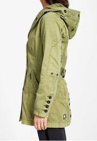 Olivgrön jacka med huva, med knäppda ärmar, sidofickor och en figursydd siluett. Tillverkad av ett strukturerat tyg.