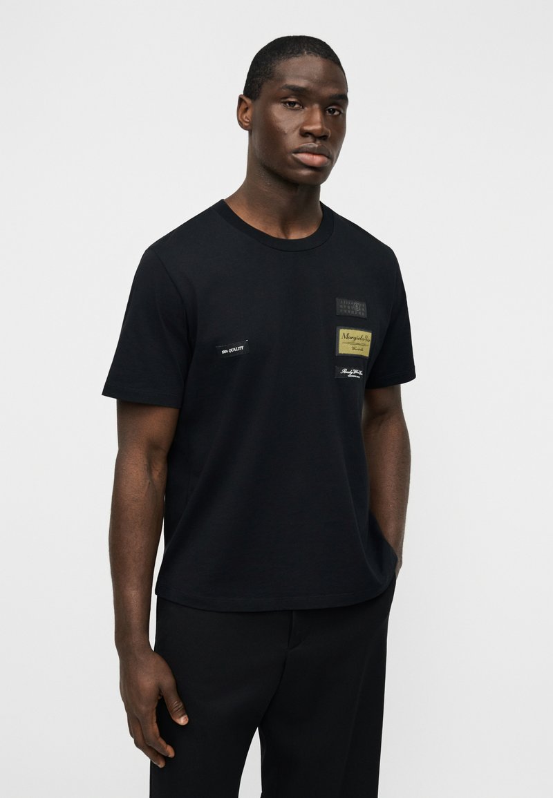 T-shirt en coton noir à manches courtes, avec plusieurs patchs en tissu beige et noir. Encolure ronde, coupe décontractée, design minimaliste.