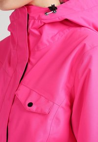 Detailní záběr na jasně růžovou bundu s vysokým límcem, černou kapsou na zápěčku a nastavitelným stahováním v oblasti krku.