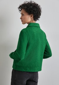 Grön fleecejacka med en texturerad yta, krage, långa ärmar och sidfickor, bärs över mörka jeans.