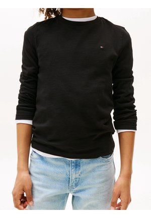 Persoon die een zwart shirt met lange mouwen draagt over een wit topje en lichtblauwe spijkerbroek, staand met armen ontspannen langs het lichaam.