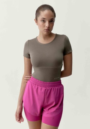 Mladá žena v hnědém tričku s krátkými rukávy a jasně růžových sportovních kraťasech s vrstvami, stojící před jednoduchým světlým pozadím.