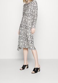 Žena v dlouhé midi šatech s černobílým vzorem a asymetrickým spodním lem, doplněná černými otevřenými mules na podpatku, před jednobarevným pozadím.