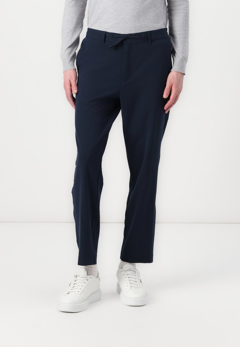 Marineblaue Hosen aus einem glatten Stoff, mit einem Kordelzugbund und einer maßgeschneiderten Passform. Kombiniert mit weißen Sneakers.