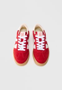 Adidași roșii din piele întoarsă, cu șireturi albe, dungi laterale și talpă de gumă. Prezintă un vârf rotunjit și o textură superioară netedă.