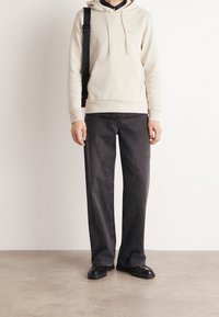 Felpa beige chiaro con tasca a marsupio, abbinata a jeans larghi grigi scuri. L'outfit presenta un design semplice ed è completato da scarpe nere.