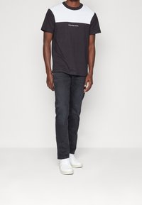 Camiseta negra con panel superior blanco, que presenta un logo de marca bordado. Combinada con jeans oscuros y zapatillas blancas, de pie contra un fondo neutro.
