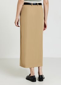 Femme portant une jupe midi beige et une ceinture noire, assortie à des mocassins brillants noirs, se tenant contre un fond blanc.