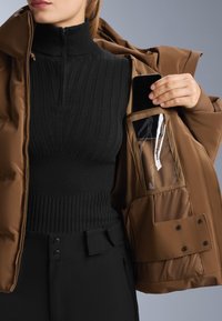 Smeđi pufer jakna s visokim ovratnikom, zatvaranjem na patentni zatvarač i unutarnjim džepom s prozirnom stranom. Crni rebrasti top ispod, slim-fit hlače.