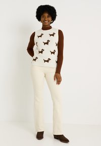 Gilet en tricot crème avec des motifs de chiens marron, superposé sur un pull à col roulé côtelé marron. Assorti à un pantalon évasé de couleur claire et des chaussures marron.