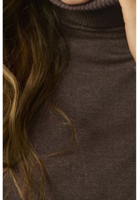 Cheveux longs bruns sur un pull à col roulé côtelé marron foncé, vue partielle du cou et de l'épaule.