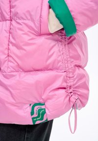 Nafouknutá růžová bunda se zelenými akcenty, elastickým stahovacím šňůrem na spodním okraji a štítkem s vlnkovým designem na boku. Hladká, lesklá textura.
