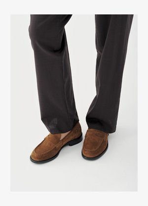 Mocassins en daim marron avec une texture lisse, ornés de détails cousus à l'avant et une semelle en caoutchouc noire, associés à un pantalon noir.
