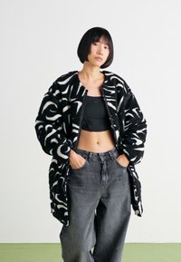 Mikina z fleece s oversize strihom a čierno-bielym vzorom s textúrovaným povrchom, v kombinácii s čiernym crop topom a vysokými šedými džínsami.